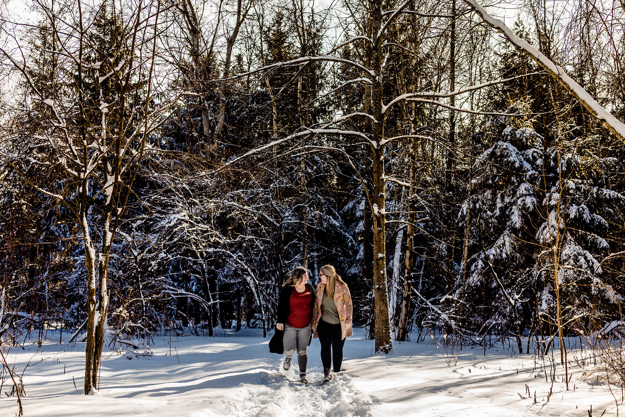 Engaged couple hold hands walking together down a snowy path in a winter forest.