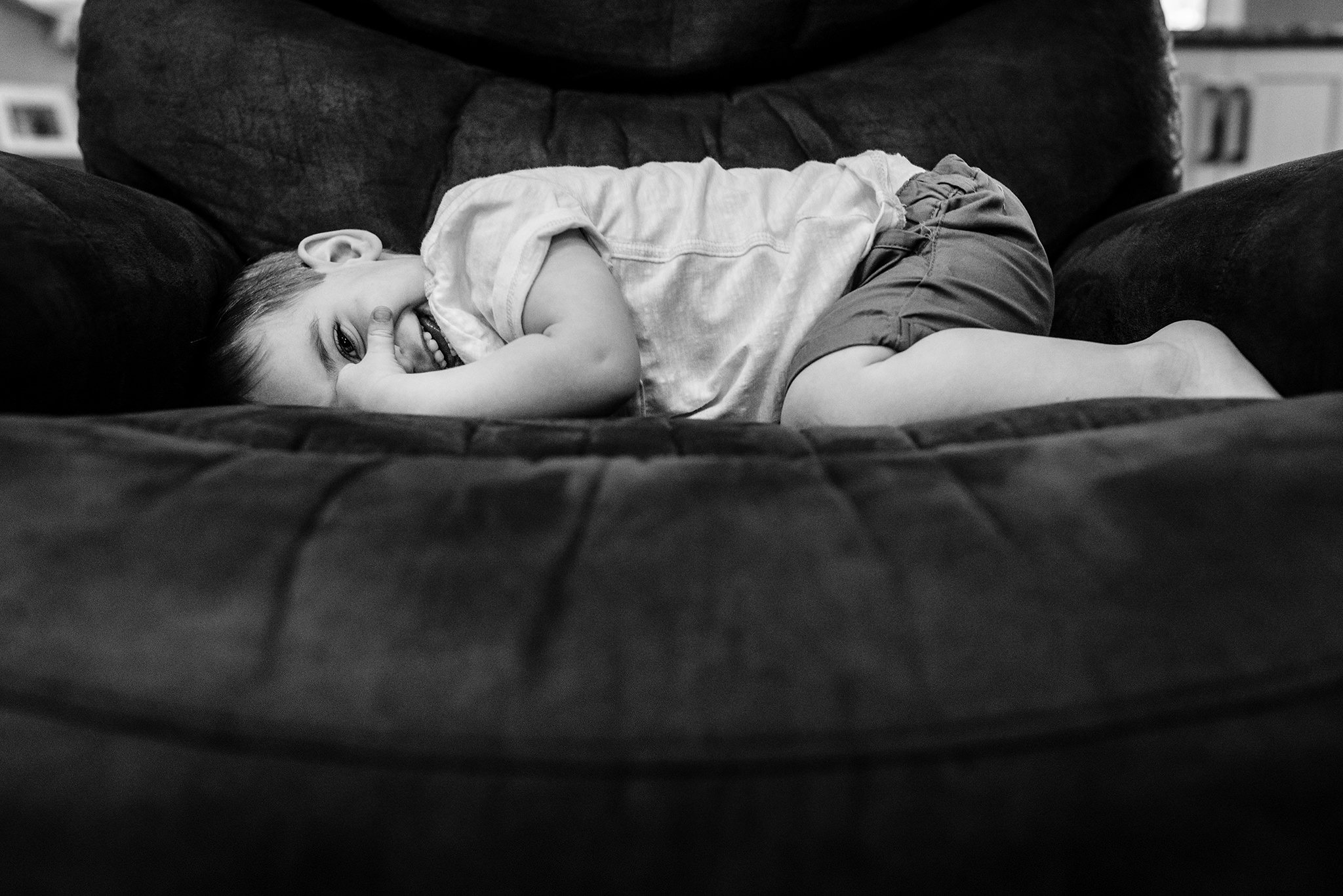 Young boy smiles while laying across a recliner chair with his arms and legs tucked in.