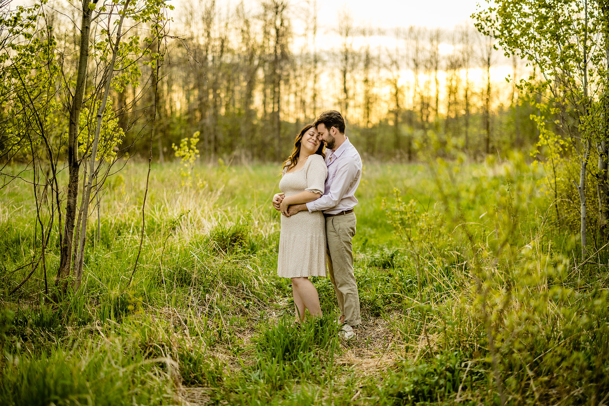 Man hugs his fiancee from behind as she smiles back at him in a green field by a forest.