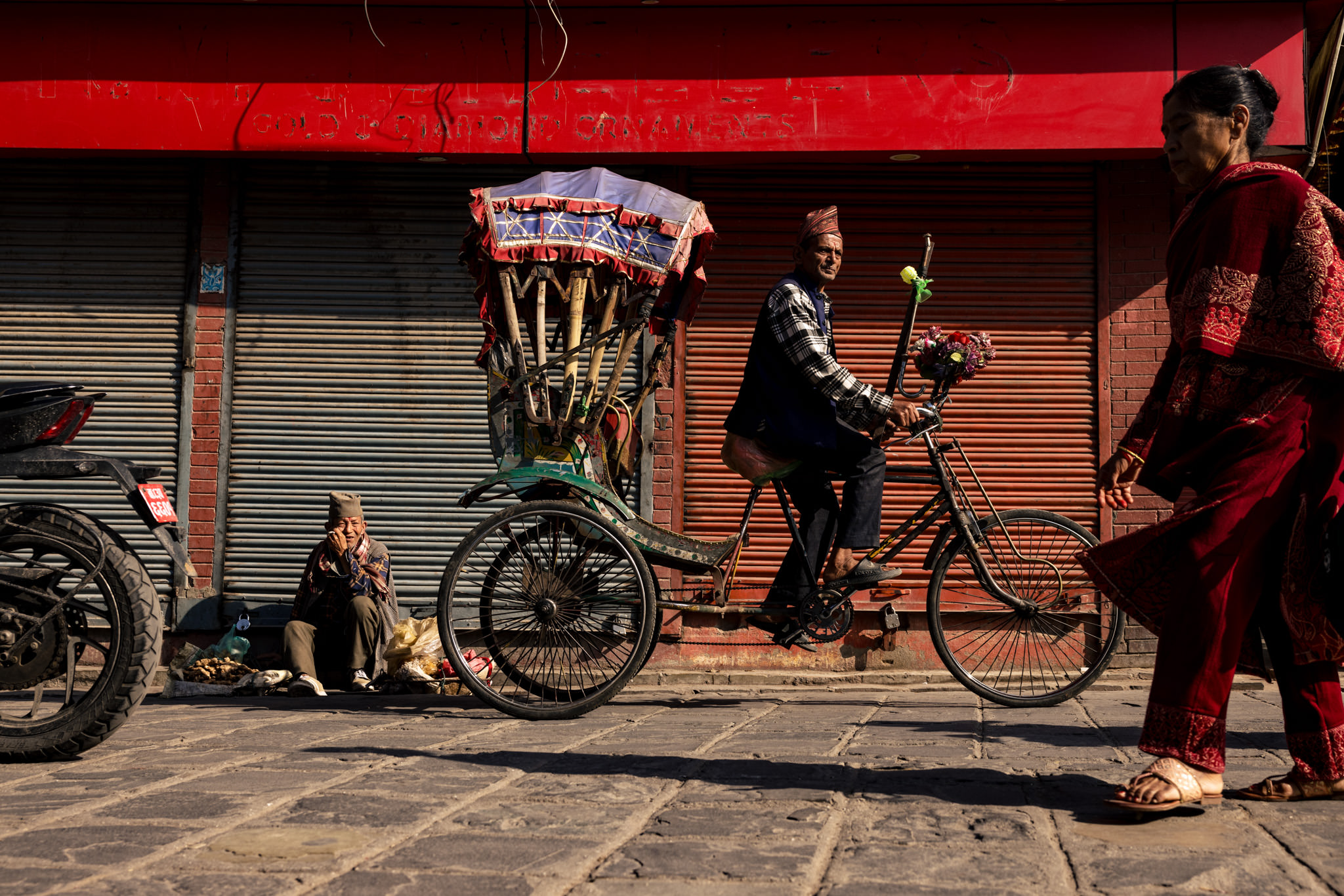 Nepali man squats by the curb with produce as autorickshaw passes in streets of Kathmandu